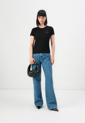 Jeune femme portant un t-shirt noir de GAP, un large jean bleu, des chaussures noires à talons pointus, tenant un petit sac à main en cuir noir sur un fond uni.