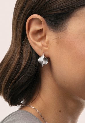 Pendientes plateados con diseño en espiral, textura suave, con una forma curva que refleja la luz, usados en la oreja de una modelo.