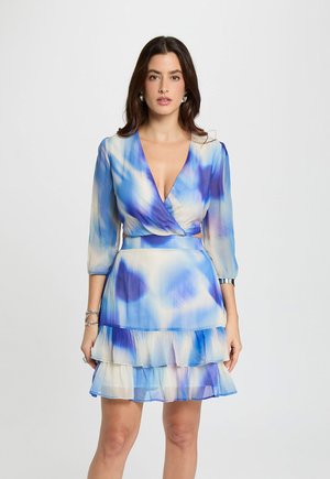 Vrouw draagt een blauw-witte tie-dye wikkeljurk met driekwart mouwen en een gerimpelde zoom, staand tegen een effen lichte achtergrond.