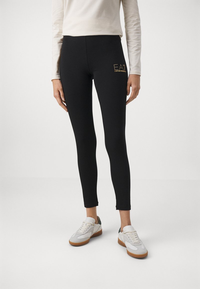 Zwarte leggings van rekbaar materiaal, met een gouden logo, hoge taille en een strakke pasvorm, gecombineerd met witte sneakers.