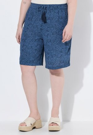 Osoba nosząca niebieskie krótkie spodenki z wzorem do kolan z troczkiem i beżowe sandały na platformie, stojąca przy białej ścianie.