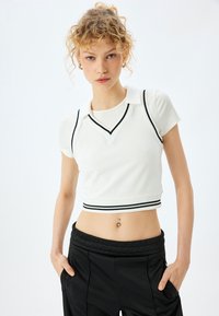 Witte crop top met korte mouwen, voorzien van een kraag, V-halslijn en zwarte gestreepte accenten langs de randen. Glad stoftextuur.