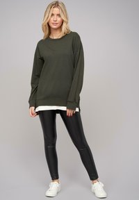 Haut à manches longues vert olive avec une coupe décontractée, superposé sur une chemise blanche ; associé à des leggings noirs et des baskets blanches. Texture lisse.