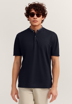 Junger Mann trägt ein dunkelnavyfarbenes kurzärmeliges Poloshirt mit geknöpftem Kragen und Sonnenbrille, steht vor einem schlichten hellen Hintergrund.