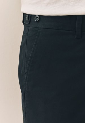 Pantalon en coton noir à texture lisse, doté de deux boutons noirs à la taille et de poches latérales pour plus de praticité.