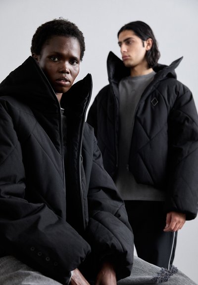 Des doudounes noires matelassées avec des cols hauts, présentant un tissu texturé et des coutures visibles. Un mannequin assis, un autre derrière.