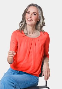 Korallfarbene Bluse mit Rundhalsausschnitt und gefälteltem Detail, kombiniert mit hellblauen weit geschnittenen Jeans. Das Model sitzt auf einem schwarzen Stuhl vor einem weißen Hintergrund.