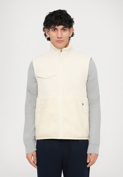 Gilet en polaire crème et beige avec une section inférieure texturée, fermeture éclair sur le devant et poche poitrine, porté sur un haut à manches longues gris.