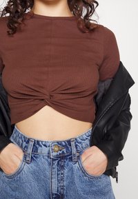 Hnědý žebrovaný crop top s twistem vpředu, kombinovaný s vysokými modrými džíny a černou koženou bundou.