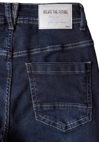 Jeans de mezclilla azul oscuro con un bolsillo trasero, detalles de costura blanca y una etiqueta de tela que dice "CREA EL FUTURO" en la cintura.