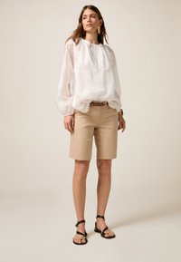 Blusa de algodón blanca con bordado, corte suelto y escote fruncido. Pantalones cortos beige de corte entallado, hasta la rodilla, y sandalias planas negras con hebillas.