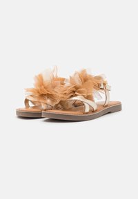 Płaskie sandały z metalicznymi paskami i dużymi beżowymi dekoracjami z tiulu w kształcie kwiatu na górze, na białym tle.