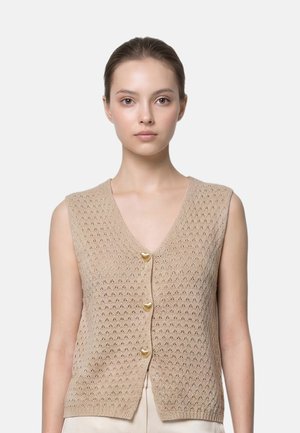 Giovane donna che indossa un gilet beige lavorato a maglia senza maniche con tre bottoni dorati a forma di cuore, in piedi davanti a uno sfondo bianco semplice.