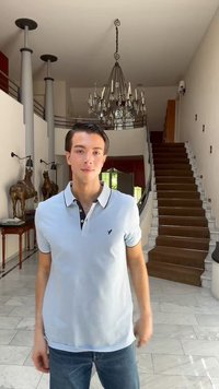 Giovane uomo con polo azzurro chiaro e jeans che si trova in una stanza con pavimento in marmo, scala, lampadario e statue di cavalli su un tavolo di legno.