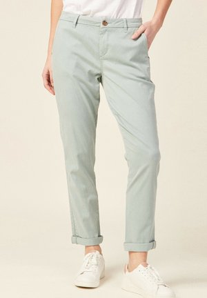 Vrouw die lichtgroene opgerolde chinos en witte sneakers draagt, staand met één hand in de zak tegen een effen achtergrond.