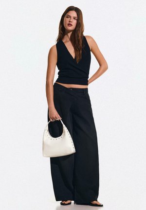 Femme debout tenant un sac à main blanc clouté, portant un haut noir sans manches et un pantalon noir à jambes larges sur fond blanc.