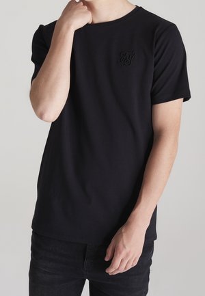 Man draagt een effen zwart T-shirt met korte mouwen en een subtiel geborduurd logo op de borst, gecombineerd met zwarte jeans, hand bij gezicht en taille.