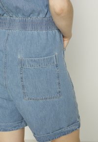 Combi-short en denim avec une coupe ajustée, taille élastique, manches courtes, une poche arrière unique, en bleu clair avec coutures visibles.