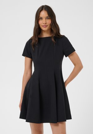 Femme aux longs cheveux bruns portant une robe noire à manches courtes avec une taille cintrée et une jupe évasée, debout devant un fond clair uni.