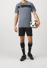 Šedý sportovní dres s černým horizontálním pruhem, černé šortky, černé fotbalové ponožky s bílými pruhy a černé kopačky držící černobílý fotbal.