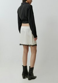 Schwarzes langärmliges Cropped-Shirt, weißer Minirock mit schwarzem Spitzenbesatz und schwarze, hochhackige Stiefeletten. Glattes Material mit strukturiertem Design.