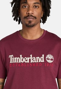 Mörkröd bomullströja med "Timberland"-logotyp i vitt och rött broderi, med året "1973" och en trädgrafik. Rund halsringning.