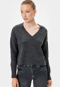Pull en tricot gris clair avec un col en V, texture côtelée et manches longues, associé à un jean gris foncé. Design simple et ajusté.