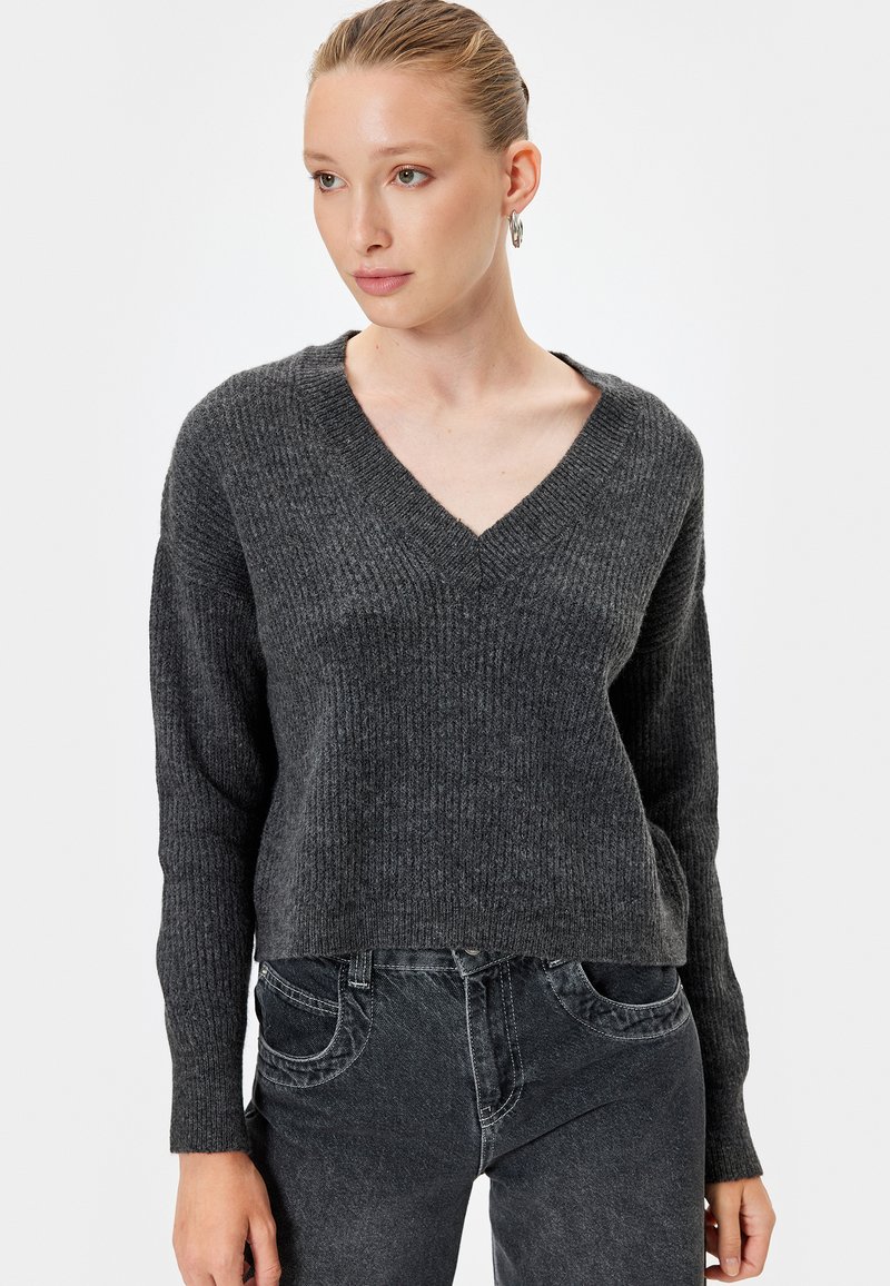 Suéter de punto gris heather con escote en V, textura acanalada y mangas largas, combinado con jeans gris oscuro. Diseño simple y ajustado.