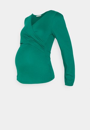 Grünes, langärmliges Schwangerschaftsoberteil aus dehnbarem Stoff. Mit Wickeldesign und gerafften Details, die einem wachsenden Bauch gerecht werden.