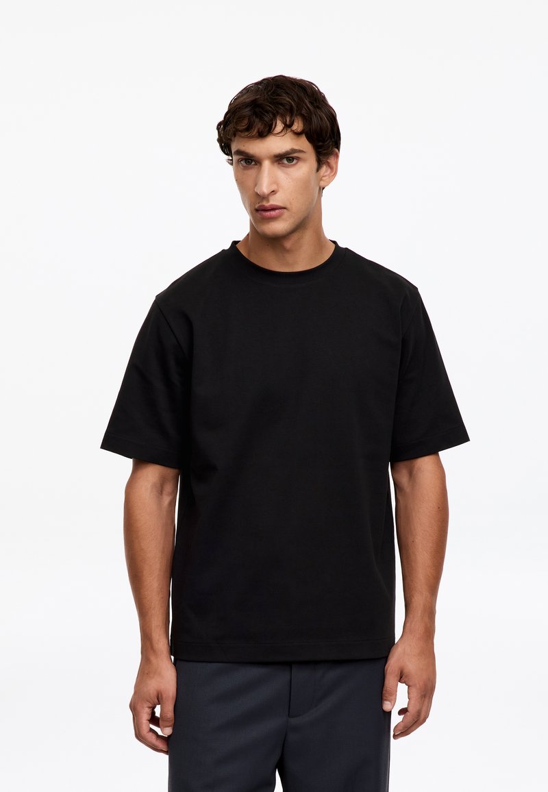 Jeune homme aux cheveux courts et bouclés portant un t-shirt noir uni à manches courtes et un pantalon sombre, debout devant un fond blanc.