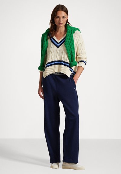 Creme kabelstrik sweatshirt med grønne accenter og v-udskæring, lagdelt med en grøn strik cardigan, stylet med marineblå brede bukser og beige sneakers.