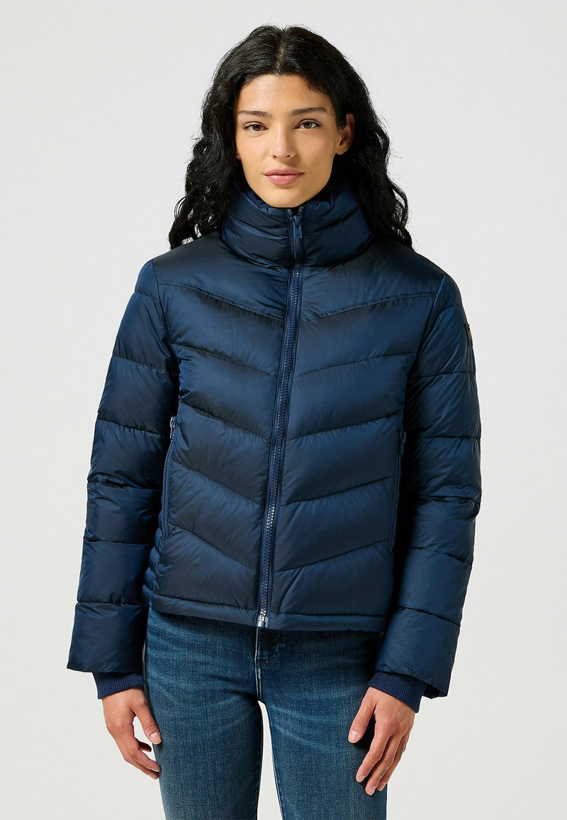 Doudoune bleu marine avec un col montant, fermeture éclair, surpiqûres en chevrons et poignets côtelés. Portée par-dessus un jean. Texture douce et lisse.