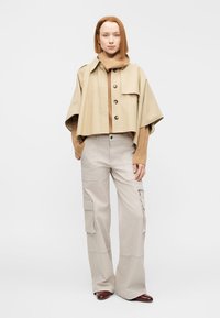 Veste beige courte avec boutons, associée à un col roulé marron et un pantalon cargo beige clair. Le mannequin pose devant un fond uni.