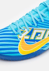 Ljusa blå sportskor med en texturerad ovandel, blå snörning och en framträdande gul Nike-swoosh med grafiska detaljer i gult och vitt.