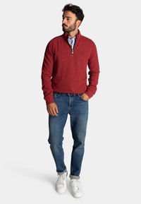 Rode zip-up sweater met een hoge kraag, gedragen over een geblokt shirt, gecombineerd met blauwe denim jeans en witte sneakers. Casual pasvorm en textuur.