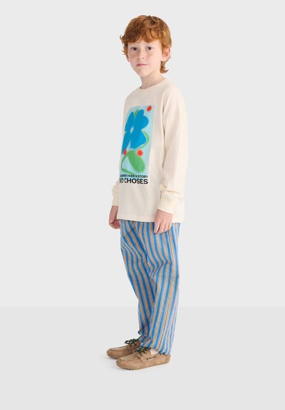 Enfant aux cheveux roux portant un sweat-shirt crème avec un motif de fleur bleue et du texte, un pantalon rayé bleu et orange, et des chaussures beige, debout de profil.
