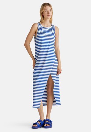 Femme dans une robe midi sans manches à rayures bleu et blanche avec une fente à l'avant, portant des sandales bleues, se tenant avec une main sur la hanche, regardant ailleurs.