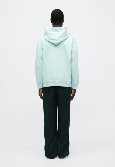 Mátově zelená mikina s kapucí na stahovací šňůrku, volným střihem a žebrovanými manžetami, doplněná širokými tmavě modrými džínovými kalhotami s zadními kapsami.