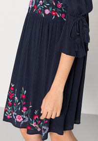 Mörkblå klänning med blommönster broderat på bysten och nederkanten, med 3/4 långa ärmar och bundna tofsdetaljer.