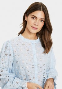 Blusa azzurro chiaro in cotone broderie anglaise, con stampe floreali, colletto arricciato e maniche a sbuffo con orli scallop.