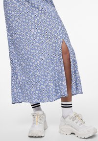 Blomstrete maxikjole i lyseblå, med sideslits og hvite tusenfryd med svarte sentrum. Kombinert med store hvite joggesko og stripete sokker.
