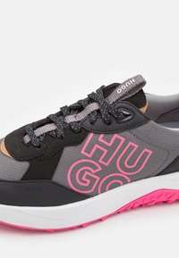 Sneaker grigia con accenti rosa, materiale nero e grigio, superficie testurizzata, logo "HUGO" in evidenza, lacci neri e suola bianca e rosa.