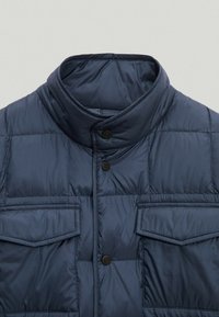 Granatowa pikowana kamizelka z wysokim kołnierzem, dwoma przednimi kieszeniami i zapięciem na zatrzaski. Wykonana z gładkiego, lekkiego materiału.