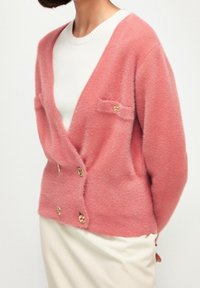 Femme portant un cardigan moelleux rose pâle avec des boutons dorés sur un pull en maille blanche et une jupe ivoire, les bras derrière le dos.