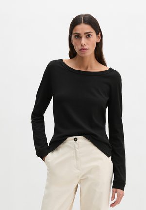 Femme aux longs cheveux foncés portant un haut noir à manches longues et un pantalon beige clair, debout les mains dans les poches devant un fond uni.