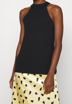 Haut noir sans manches à col halter associé à une jupe jaune pâle avec des taches noires irrégulières portée par une personne debout.