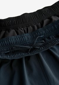 Bucșă elastică cu șnur într-un material închis, având un panou negru texturat deasupra. Material fin și ușor dedesubt cu cusături laterale.