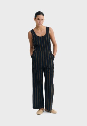 Woman standing with hands in pockets wearing a sleeveless black jumpsuit with vertical brown stripes and beige flat shoes.