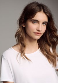 Wit T-shirt met een ontspannen pasvorm en een ronde halslijn. Zachte stoftextuur onder natuurlijk licht; model toont lang, golvend haar en neutrale make-up.