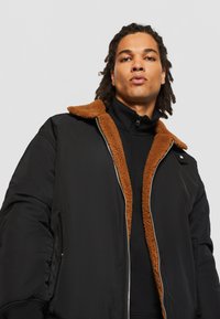 Veste bomber noire avec un col en fourrure marron, dotée d'une fermeture éclair sur le devant et de poches latérales. Le tissu semble lisse avec une finition mate.